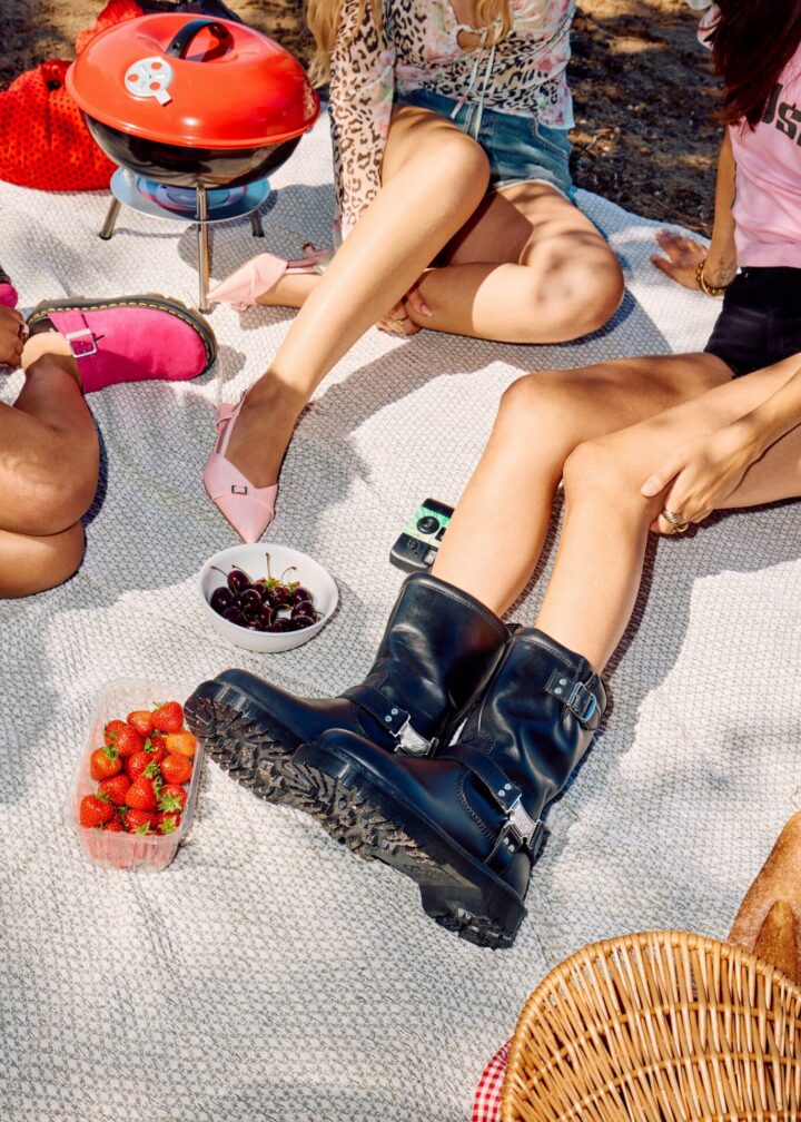 Person wearing the Dr Martens Anistone Hi in Black sitting on a picnic blanket next to to other people, one wearing a pair of pink heels and the other wearing pink Dr. Martens Zebzag mule.