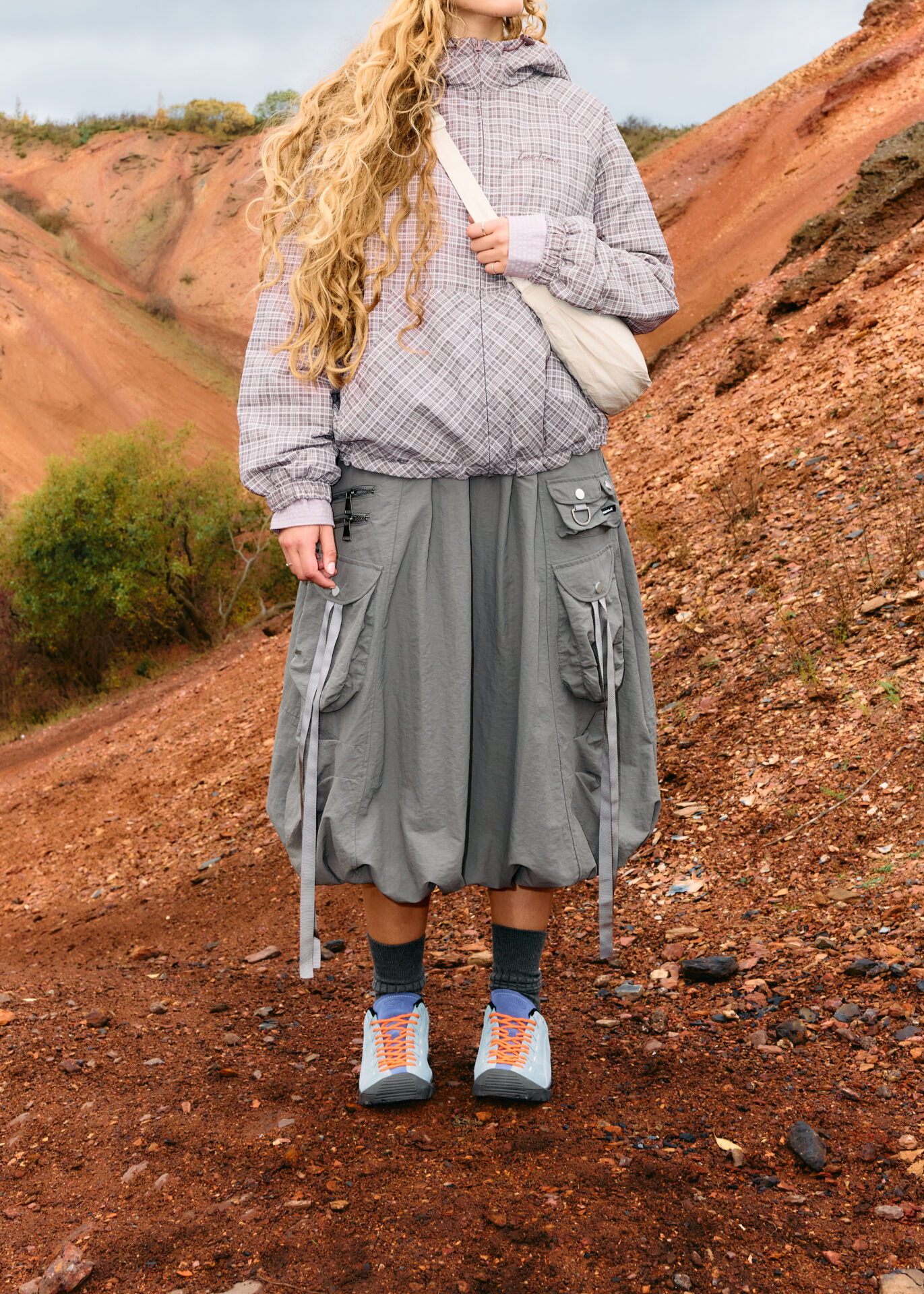 Person wearing a technical midi skirt, checked jacket, and crossbody bags with a pair of blue KEEN Jaspers.