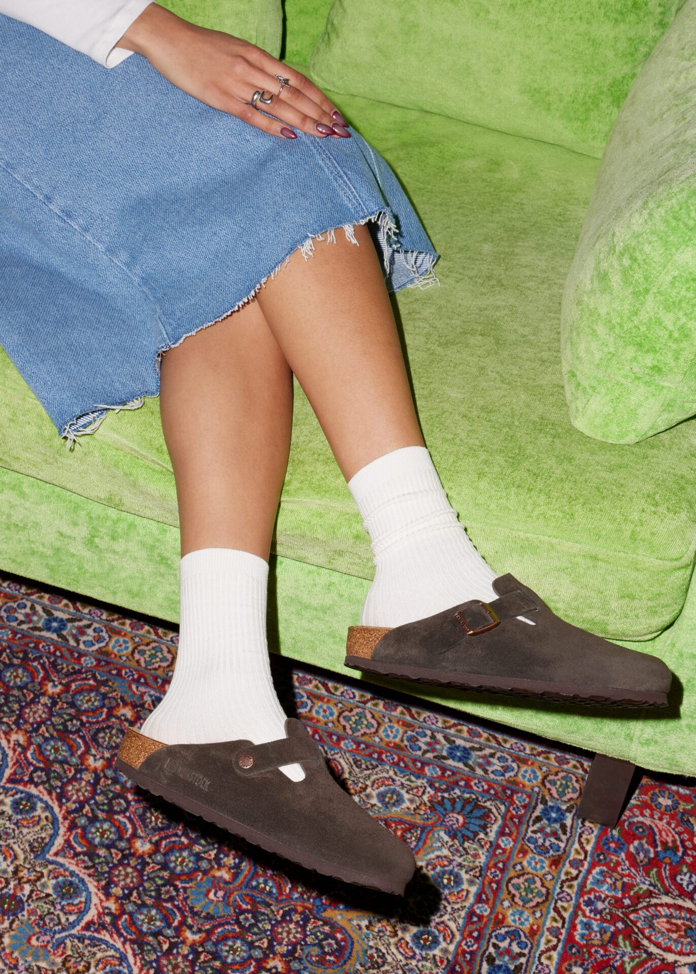 Person wearing BIRKENSTOCK Bostons in dark brown with white socks and a long denim skirt, sitting on a green sofa.