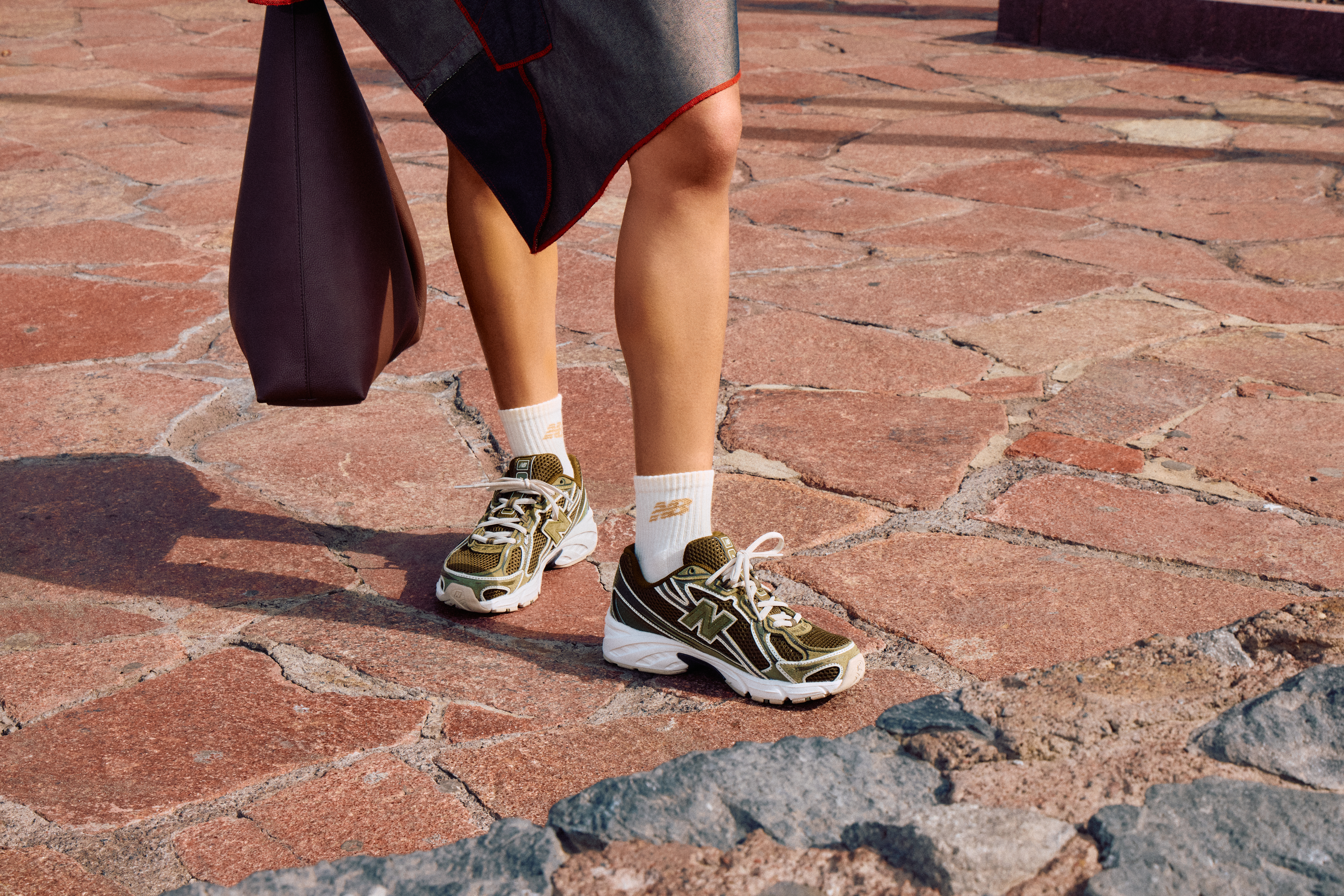 Person wearing khaki New Balance 740 with white socks and a grey asymmetric skirt, holding a brown bag.