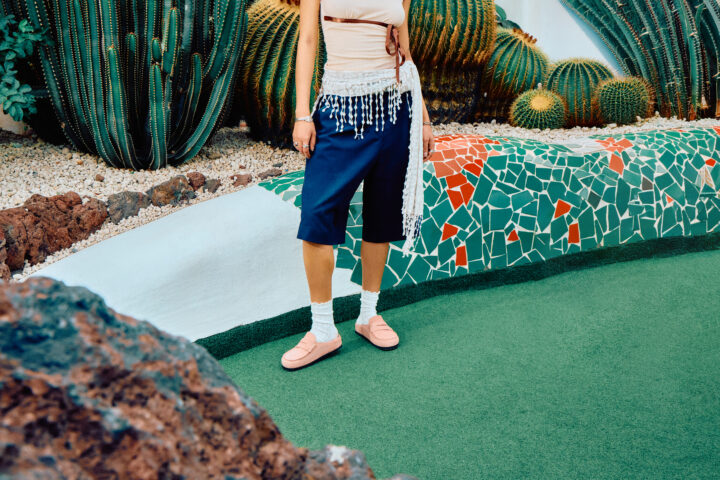 Person wearing the BIRKENSTOCK Naples in pink with white socks, long blue shorts, a peach-coloured top, and a scarf tied around their waist, standing in front of some plants.
