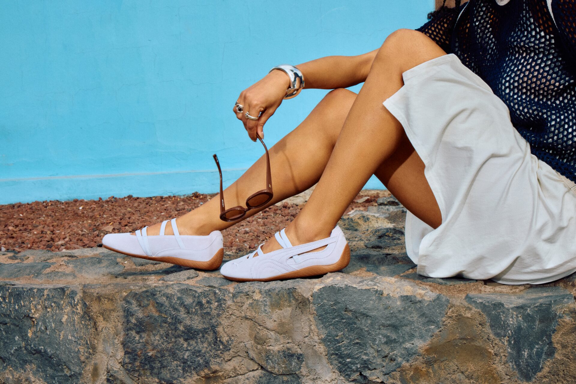 Person wearing the PUMA Speedcat Ballet in lilac with a white skirt, black netted top, and silver jewellery, holding a pair of brown sunglasses and sitting in front of a blue wall. 