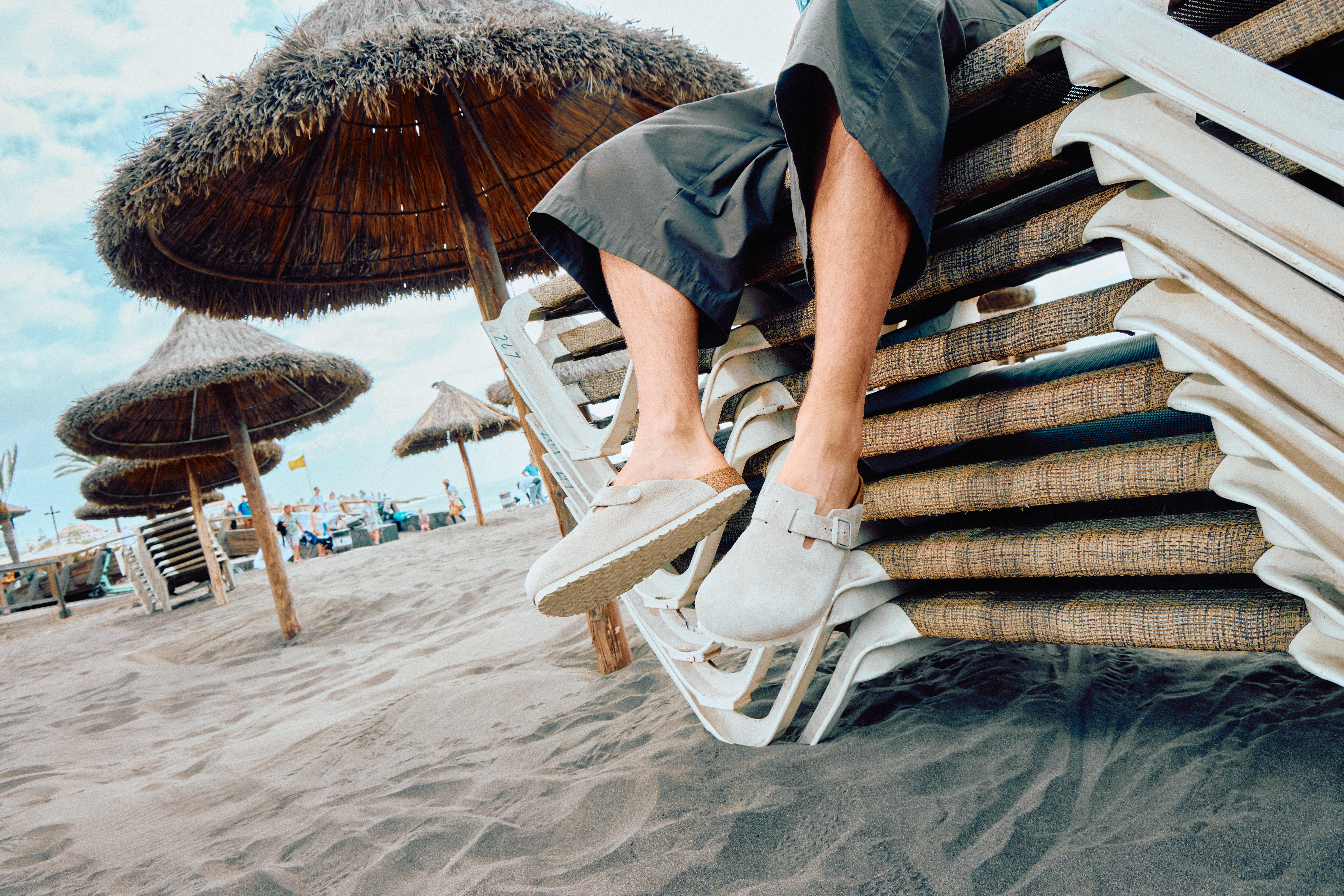 Person wearing a pair of beige BIRKENSTOCK Bostons with a pair of grey jorts, sitting on a stack of beach chairs at a beach.