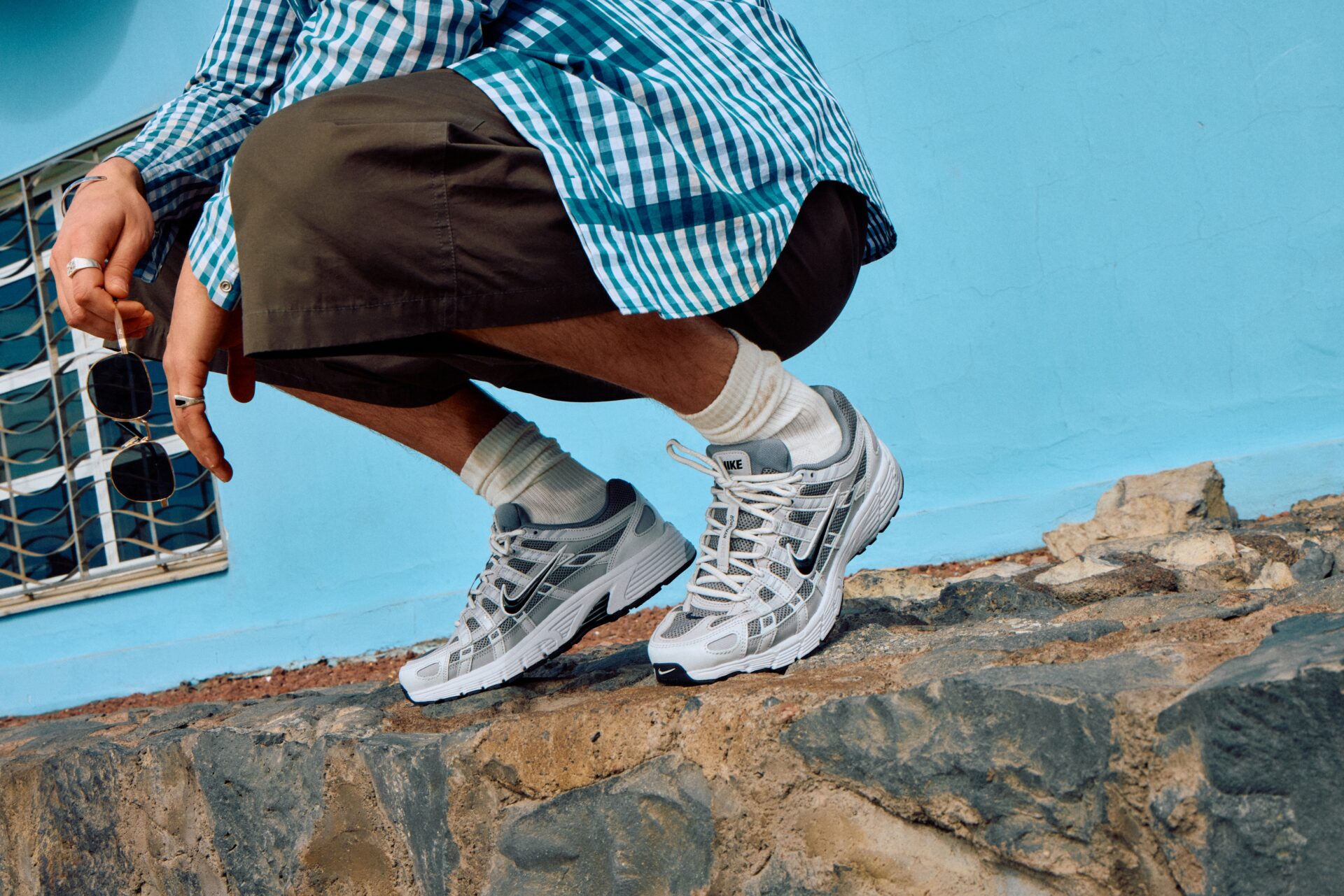 Person crouching on a brick wall wearing grey and white Nike P-6000 trainers with brown shorts and a turquoise and white checked shirt, holding sunglasses.