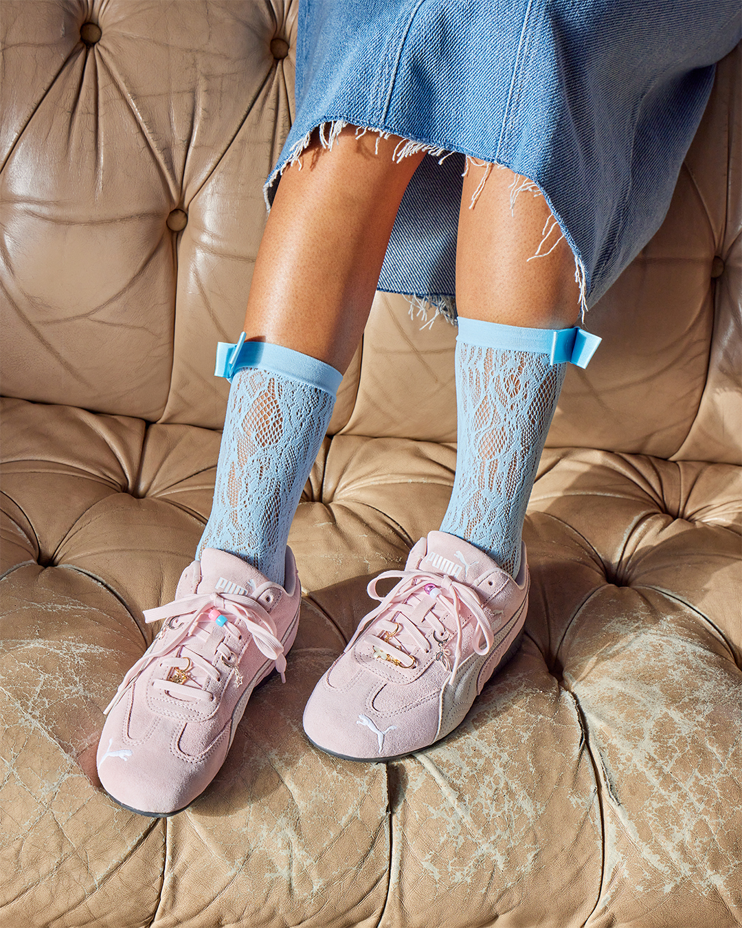 Person wearing pink PUMA Speedcat trainers with blue lace socks and a denim skirt.