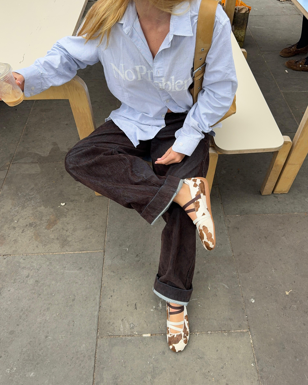 Person wearing the cow print PUMA Speedcat Ballet with jeans and a shirt, holding a coffee.