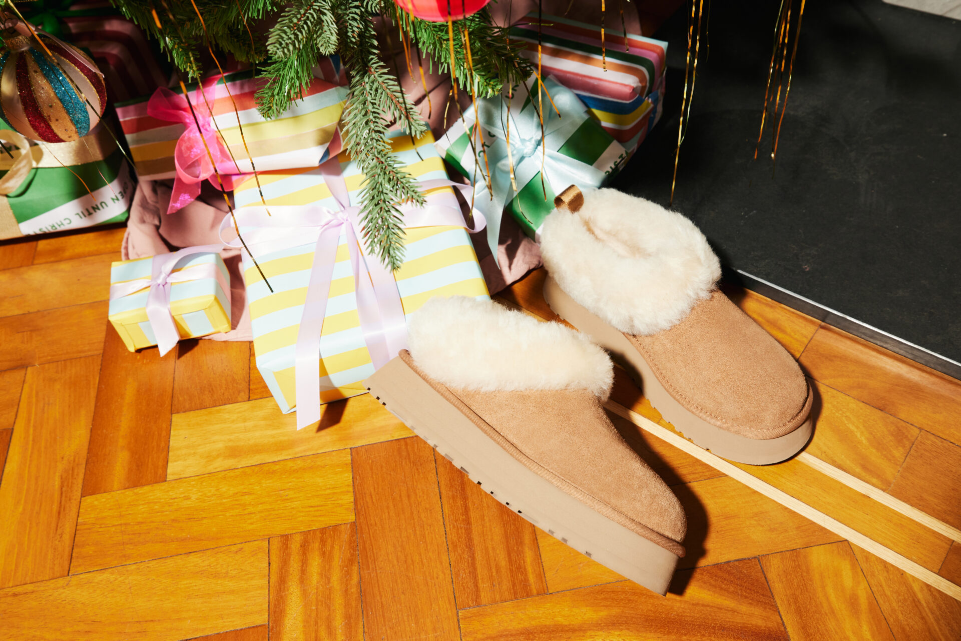 A pair of UGG Tazelles in chestnut next to other presents underneath a Christmas tree.
