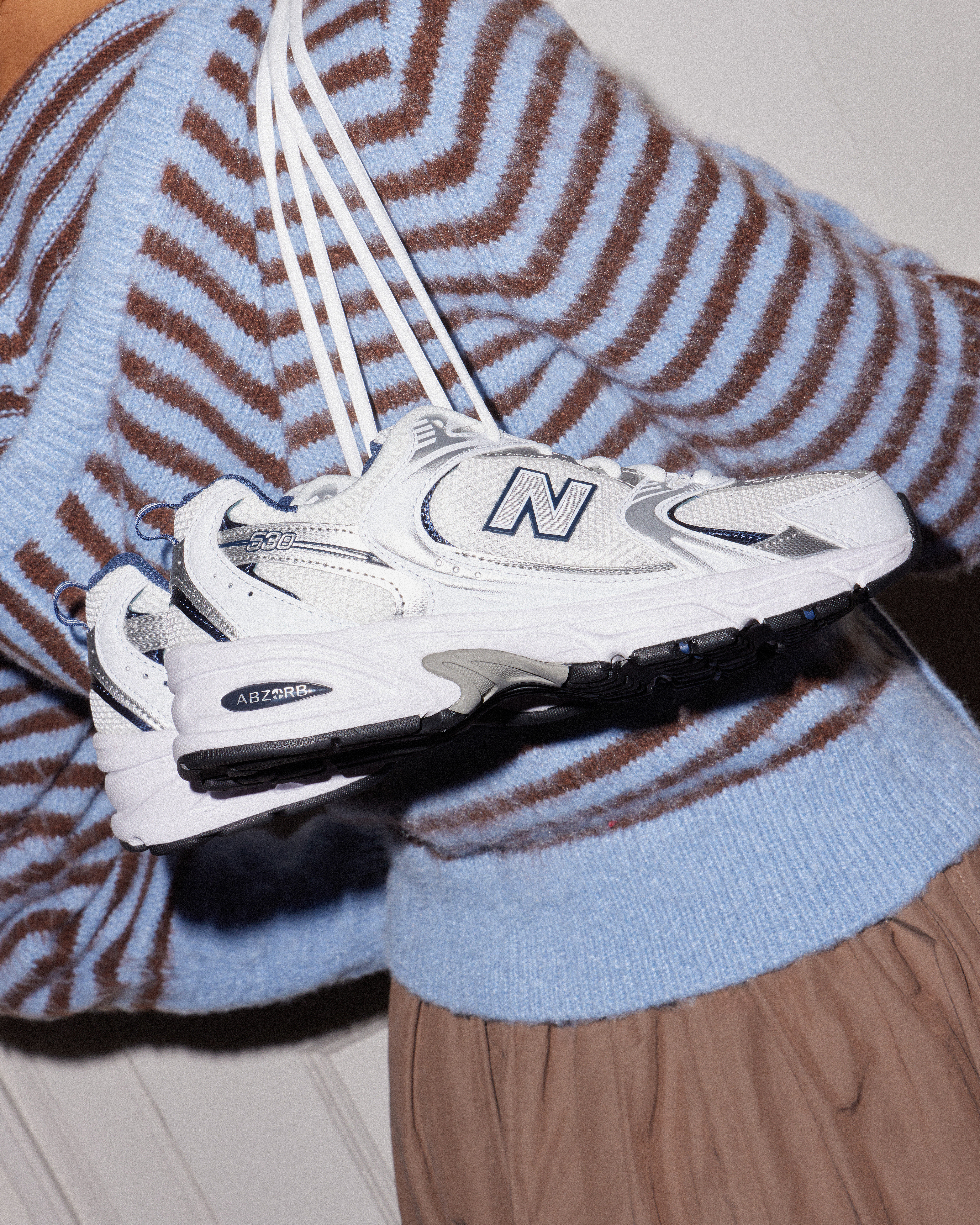 Person in a blue cardigan holding a pair of white & silver New Balance 530s.