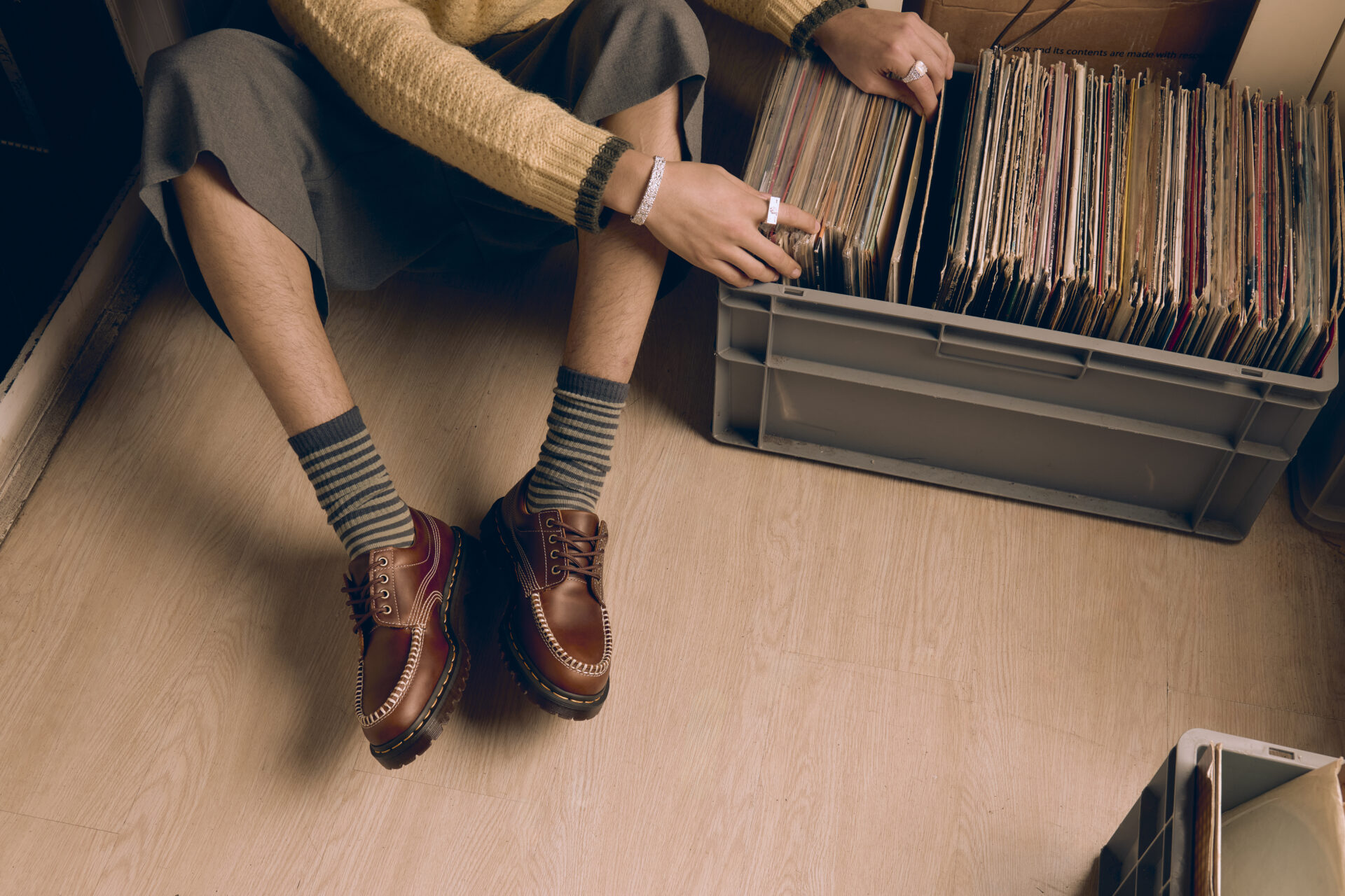 Person wearing the Dr. Martens Lowell in butterscotch brown with striped grey socks, grey shorts, and a yellow jumper while looking through a case of vinyl records.