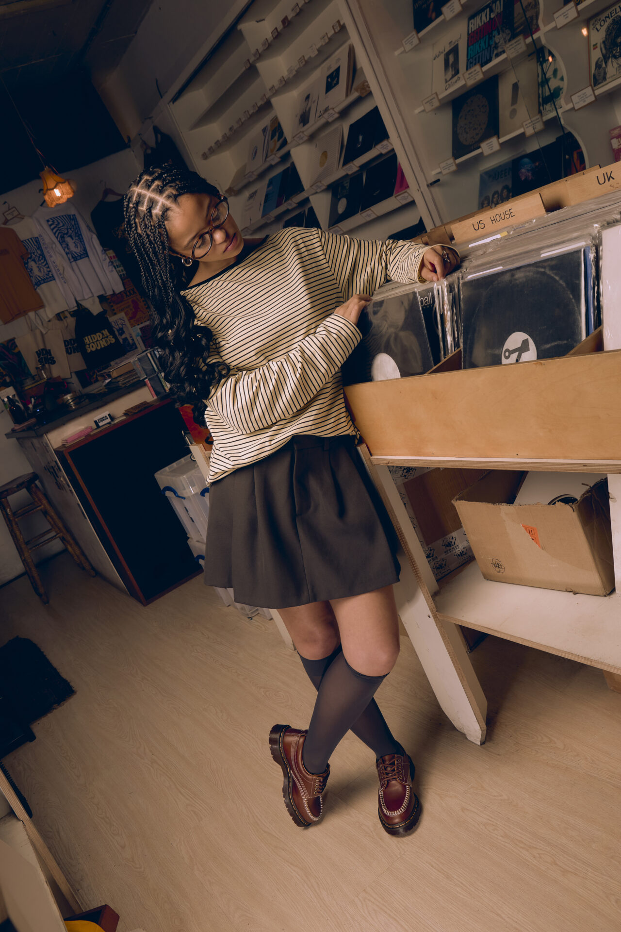 Person looking through vinyl records while wearing the cherry red Dr. Martens Lowell with grey knee high socks, a grey skirt, and long sleeved striped top.