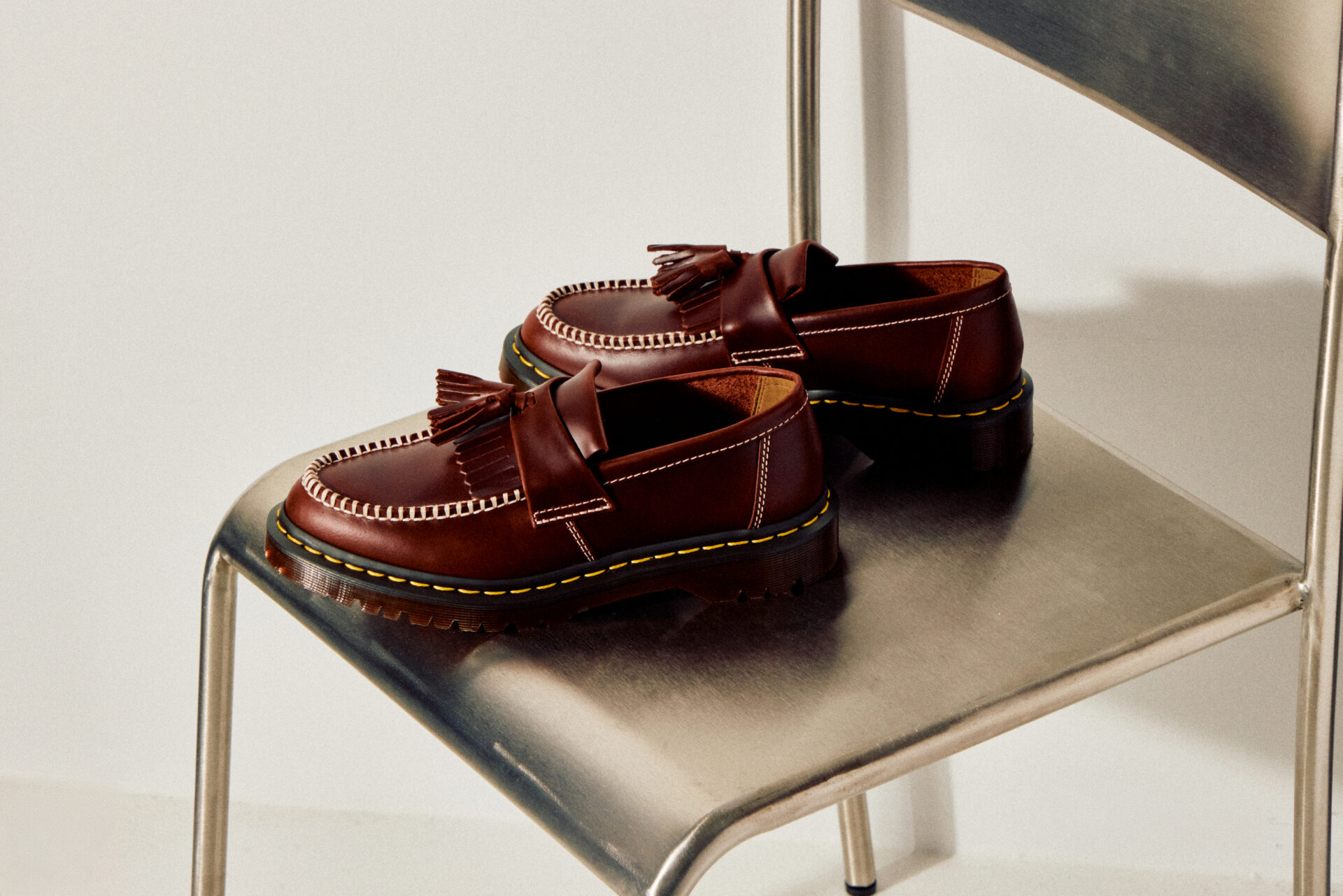 A pair of Dr Martens Adrian Ben Tassel Loafer in Burgundy sitting on a metal chair.