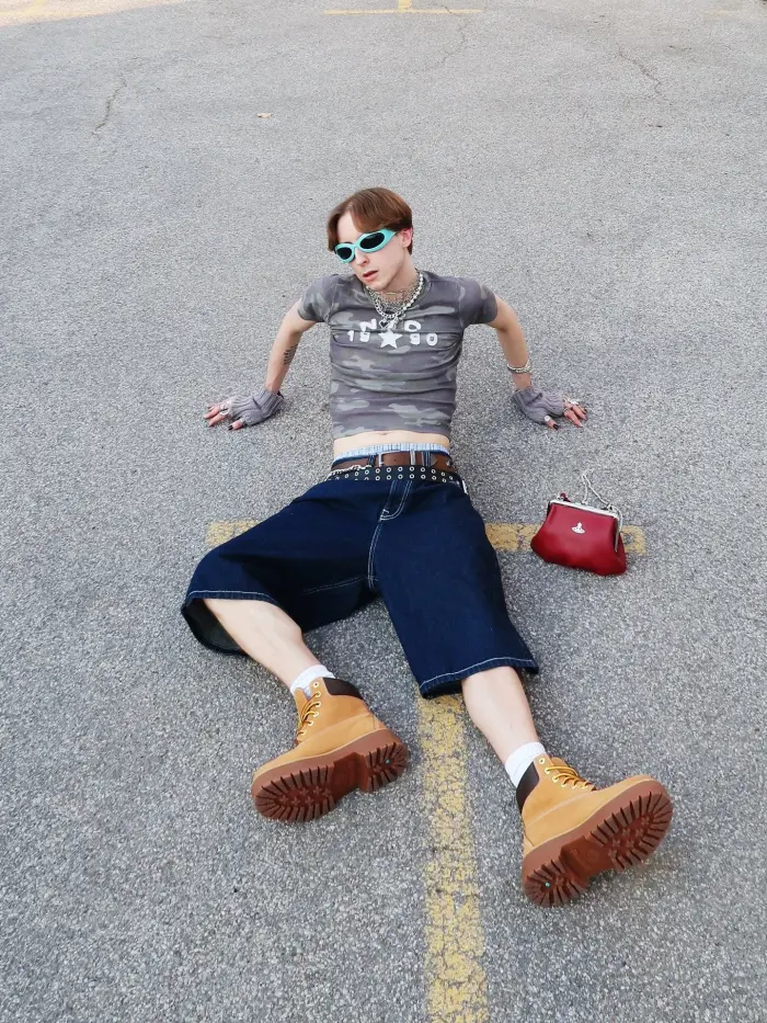 @urbanistrash (Instagram) wearing the light brown Timberland Premium 6-Inch boot with white socks, dark blue jorts, two belts, and a camo crop top, complete with grey fingerless gloves and blue sunglasses.