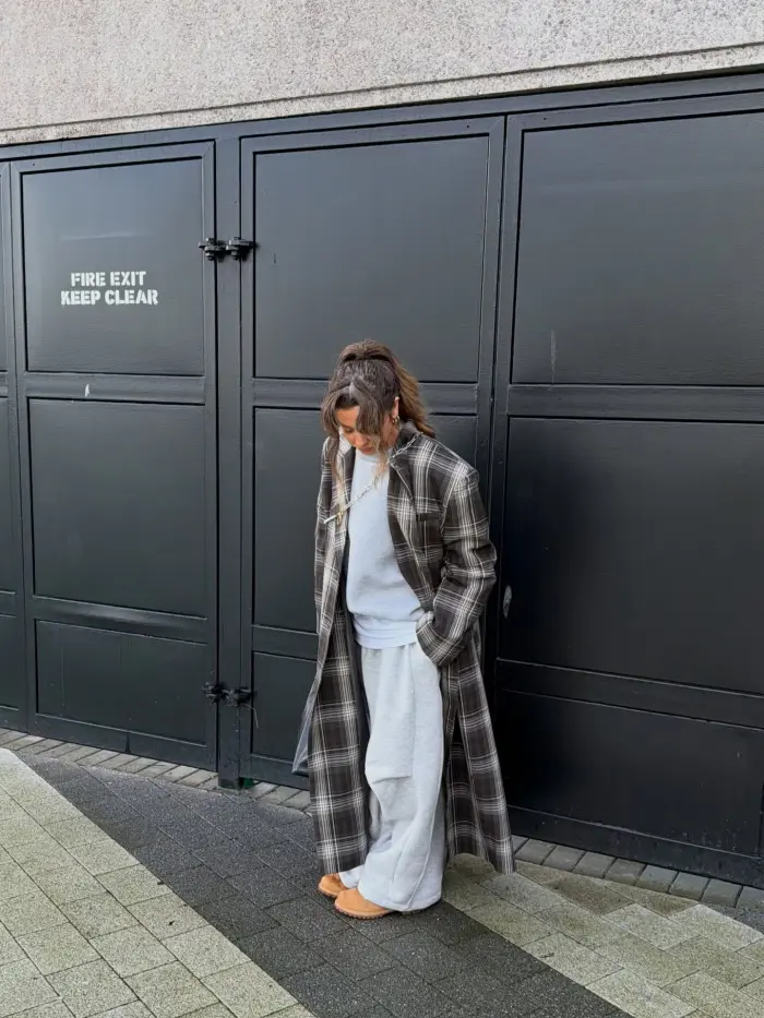 @maureenmoores_ (Instagram) wearing light brown Timberland boots with a grey jumper and grey joggers, paired with a long checked black and white coat.