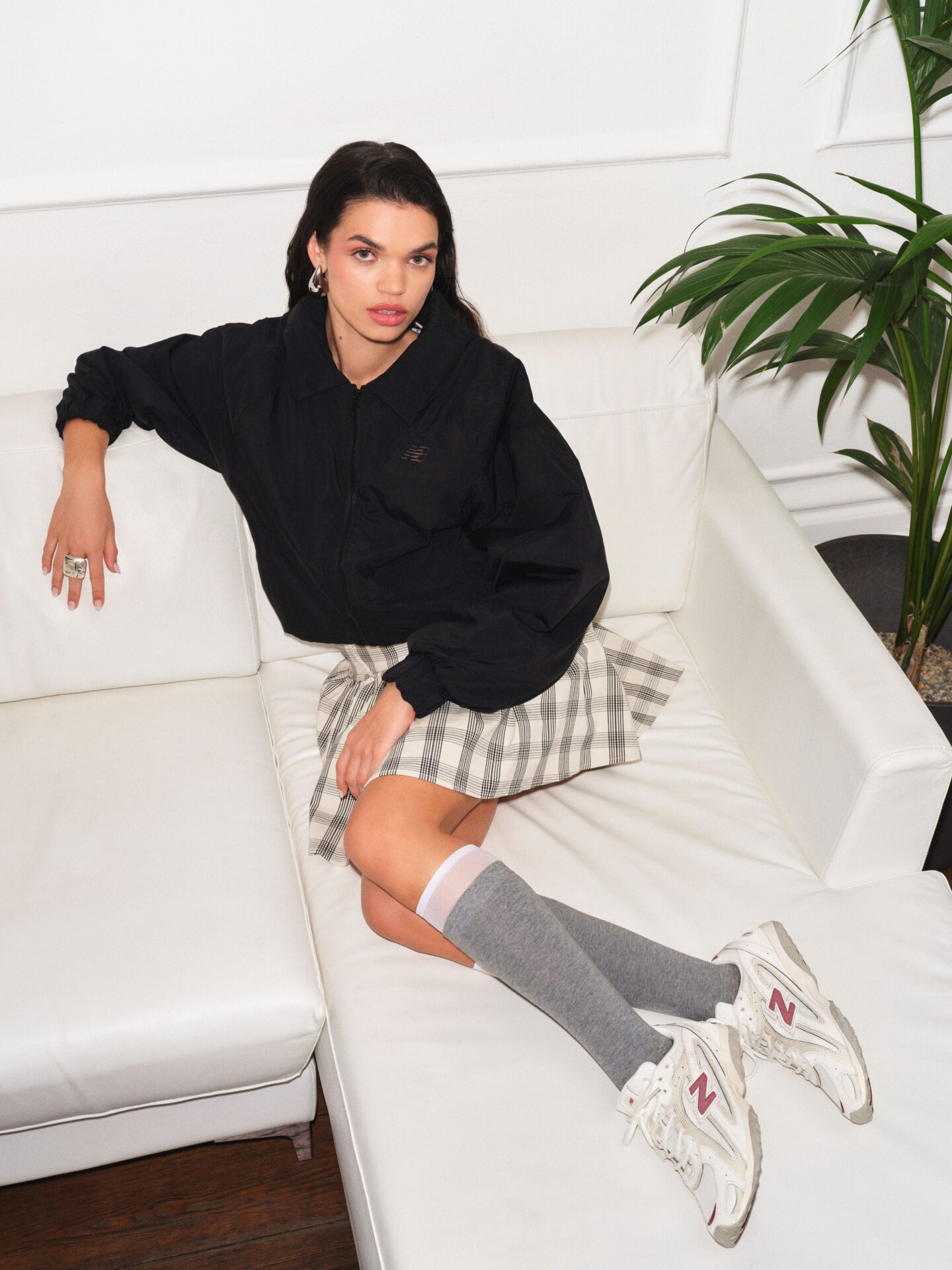 Person wearing white New Balance 204L with grey knee high socks over white mesh knee high socks, a checked white and grey skirt, black hoodie, and silver jewelry, sitting on a white couch with a plant in the background.