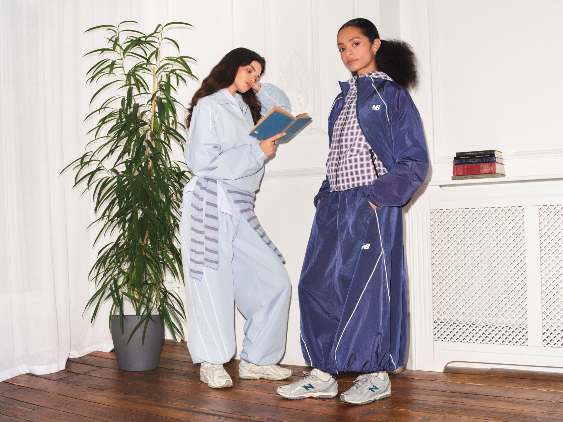 Two people wearing blue New Balance tracksuits wearing the New Balance 204L in off-white and grey & blue, one of them reading a book.