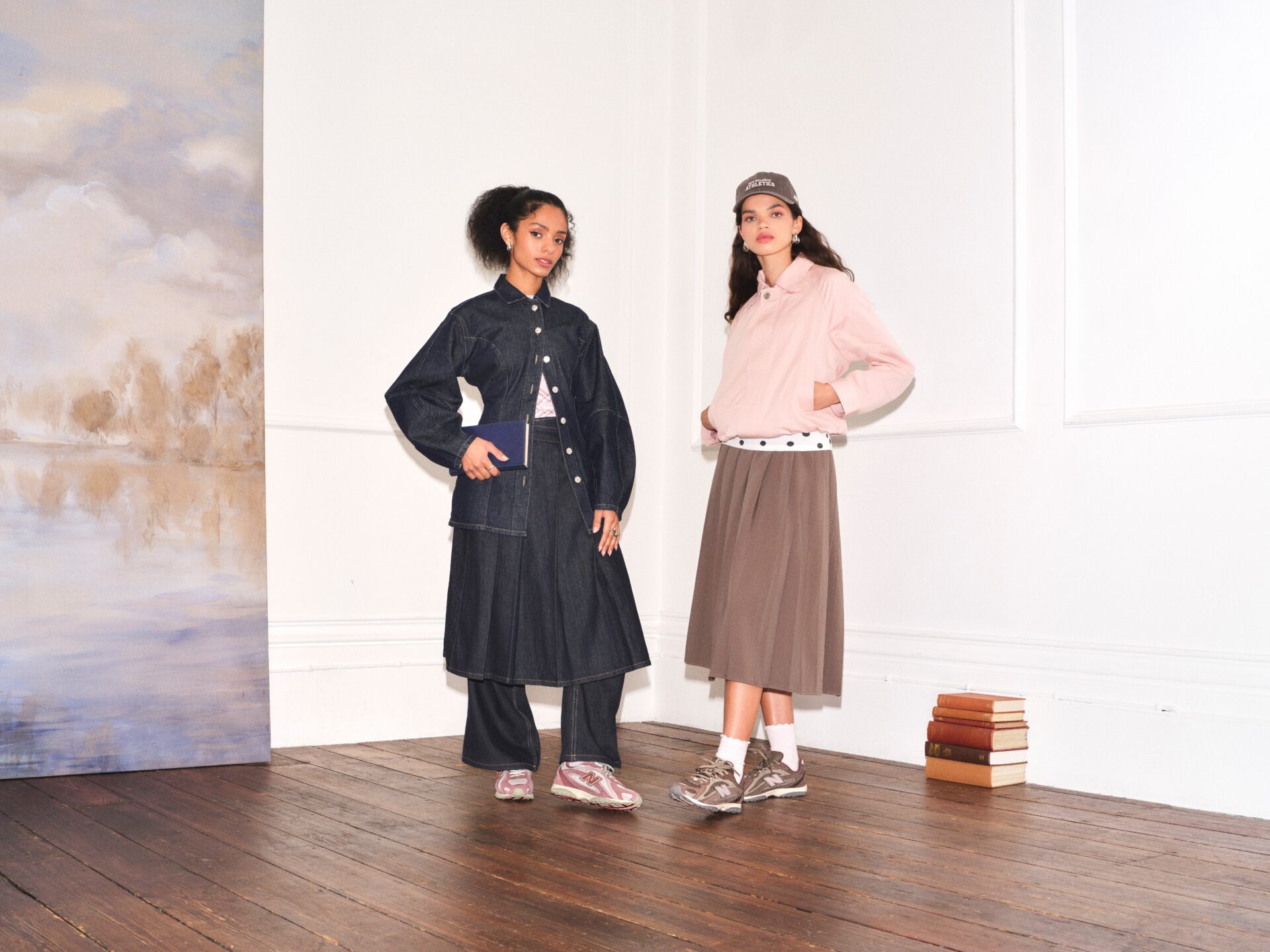 A person wearing the New Balance 204L in pink with a denim jacket, jeans, and skirt co-ord set over a white checked top, next to a person wearing New Balance 204L in brown with a brown midi skirt, polka dot top, pink jacket, and brown cap. A stack of books and a large piece of artwork are in the background.