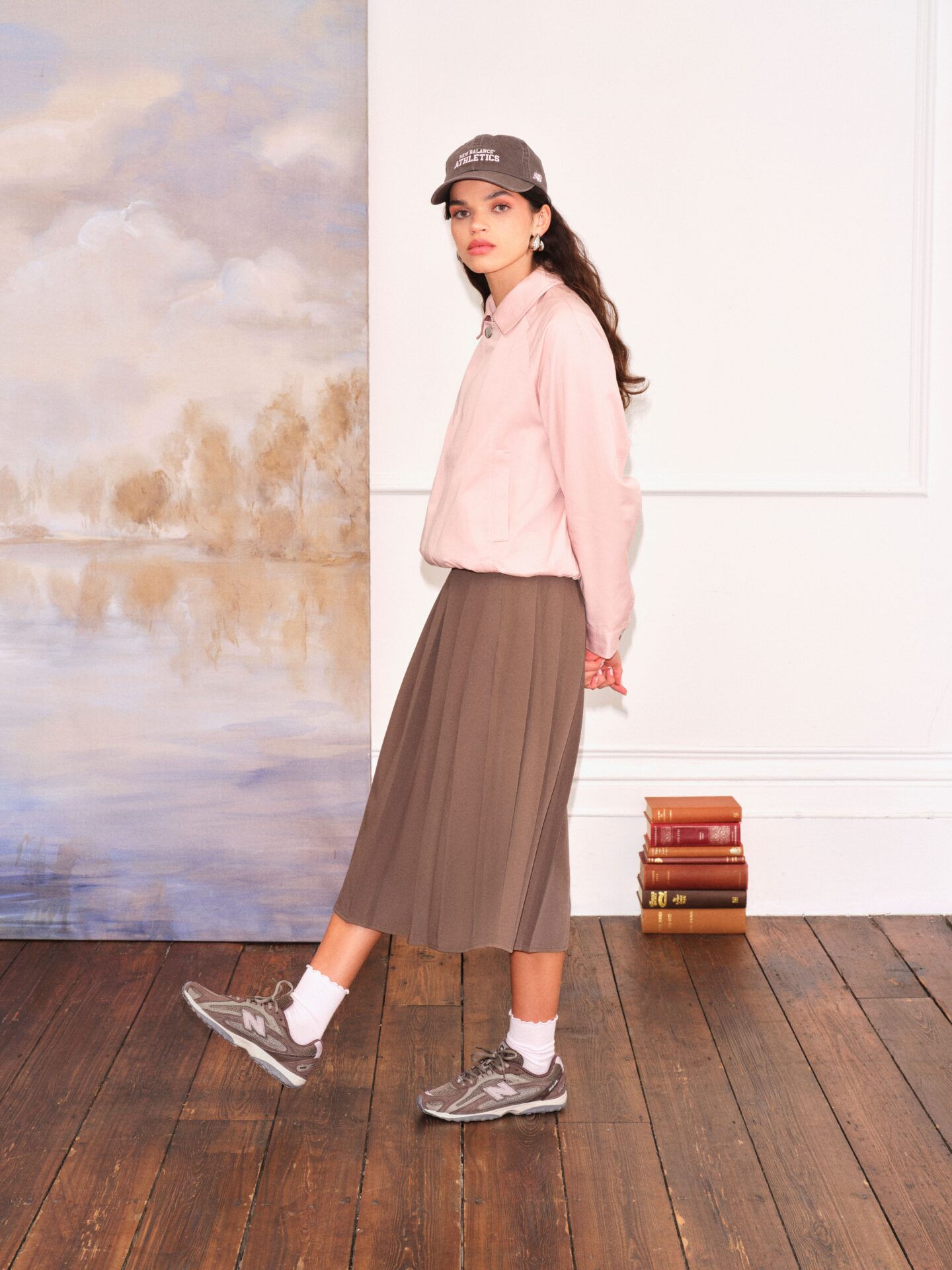 A person wearing the New Balance 204L in brown with white socks, a brown midi skirt, pink jacket, and brown shoes. A stack of books and large piece of artwork are in the background.