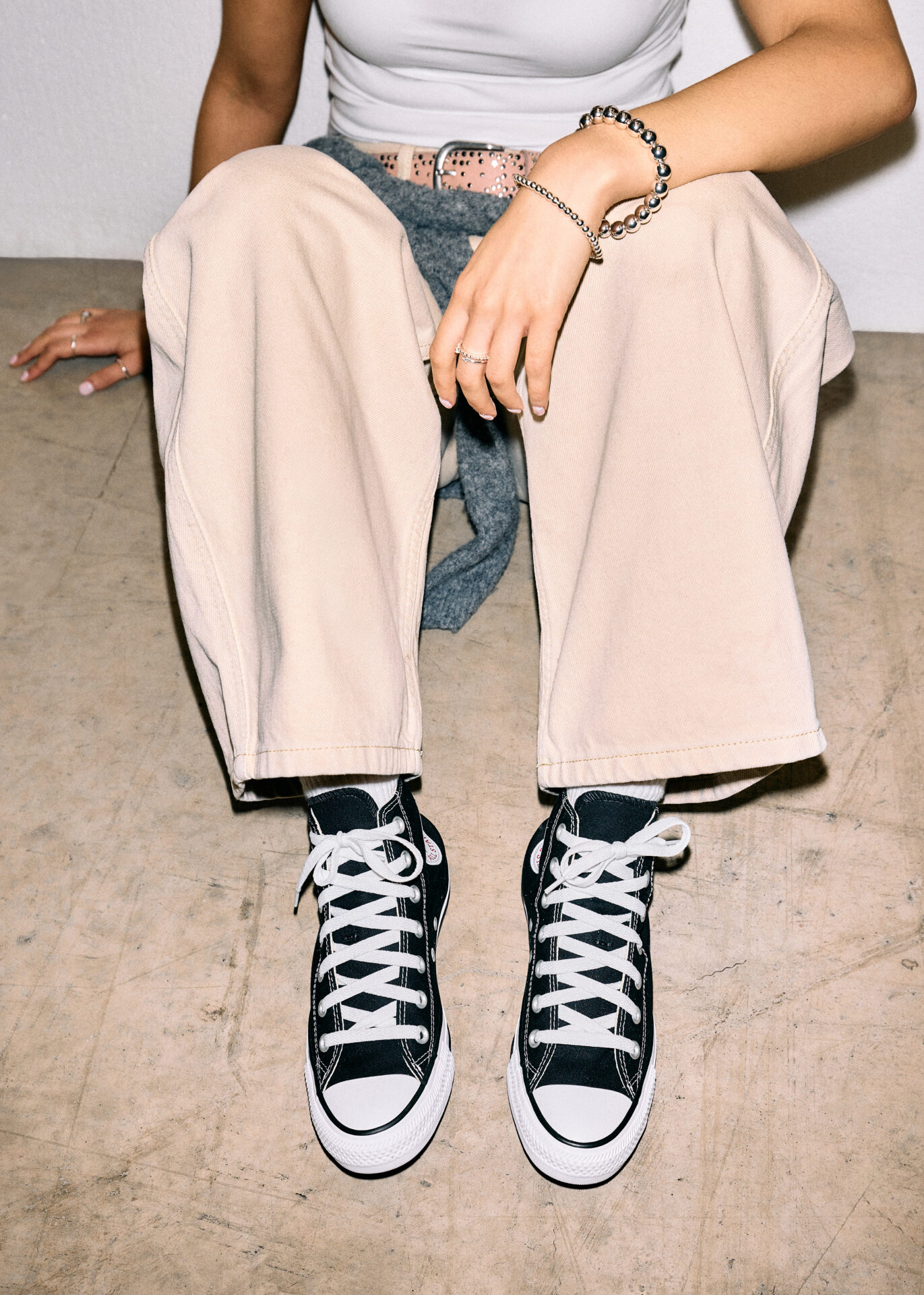 Person wearing black Converse All Star Hi with beige trousers, a white tshirt, and silver accessories.