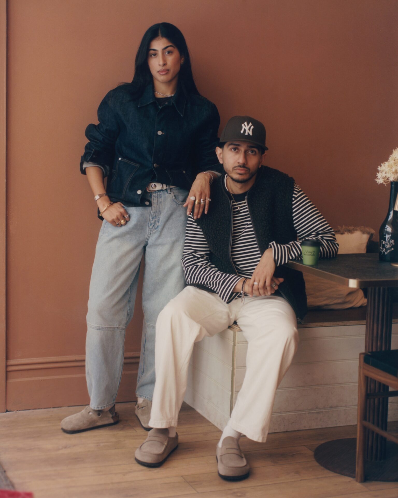 Saroop and Jasbir in their Leeds coffee and wine bar, Vinyl Grounds. Saroop wears the BIRKENSTOCK Boston in beige with light blue jeans and a dark blue jacket; Jasbir wears the BIRKENSTOCK Naples in beige with white trousers, a black and white striped tshirt, black gilet, and a cap.