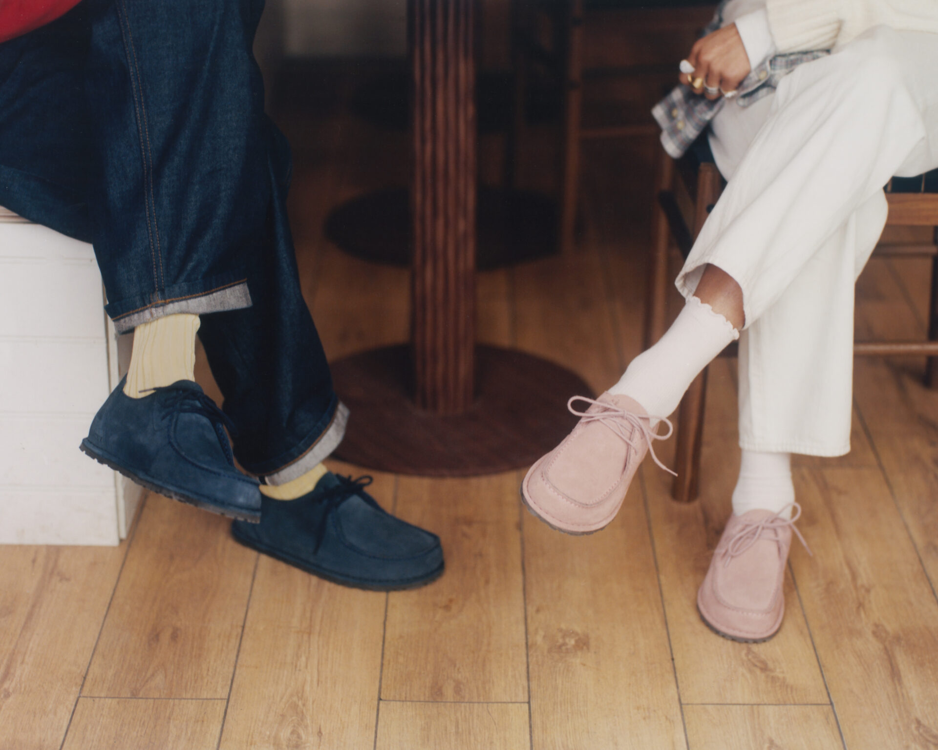 One person wearing the BIRKENSTOCK Utti in blue with yellow socks and blue jeans sitting across from another person wearing the BIRKENSTOCK Utti in pink with white socks and white trousers.