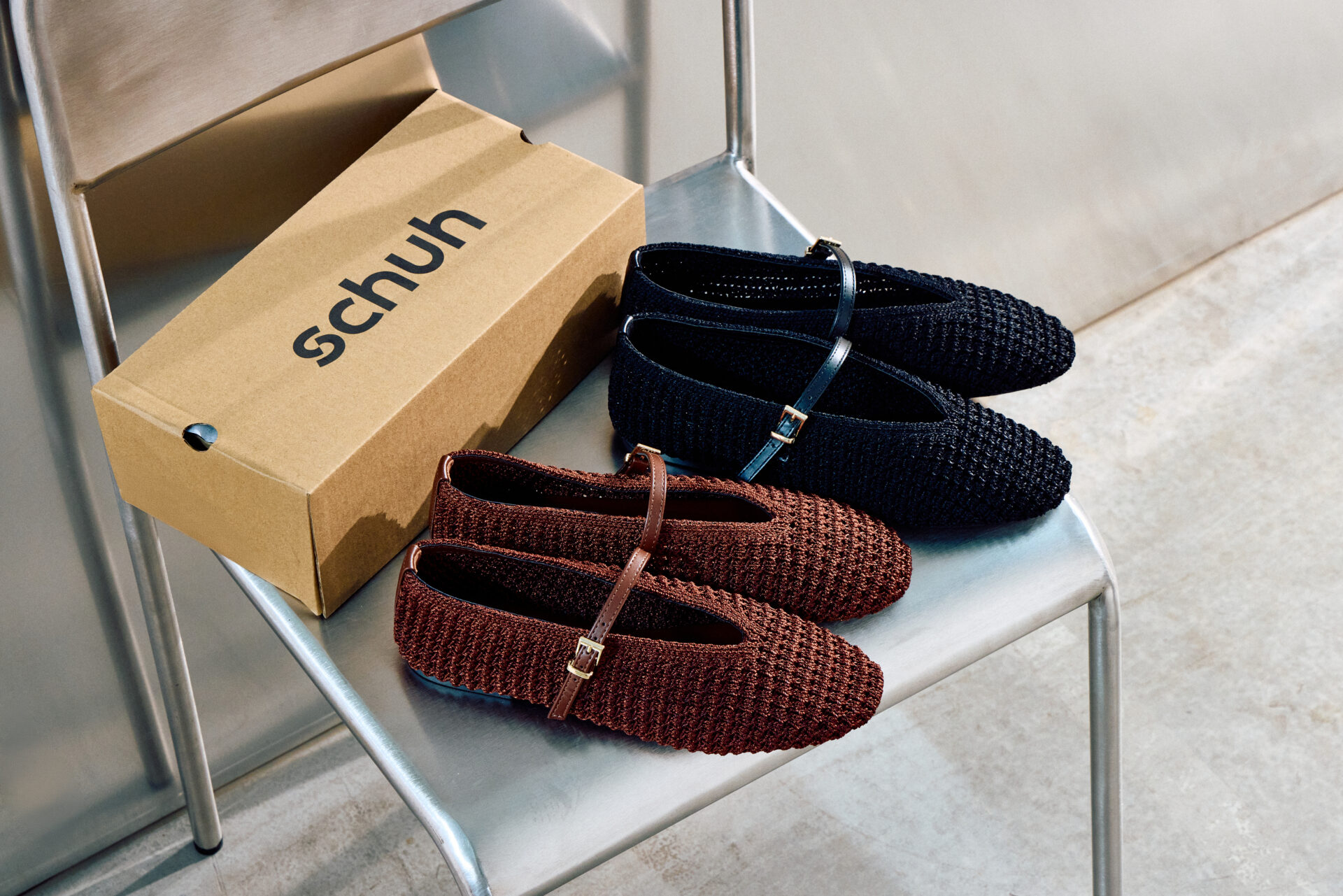 Two pairs of the schuh Lisbeth Crochet Ballerina, one in black and one in brown, sitting on a metal chair next to a schuh shoe box.