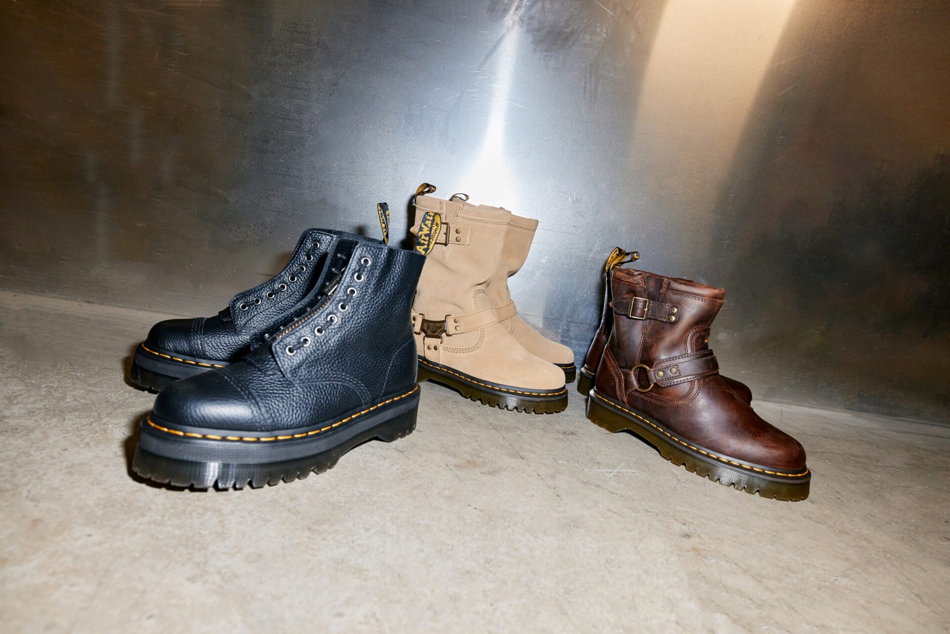 Three pairs of Dr. Martens boots - one black leather pair, one beige suede pair, and one dark brown leather pair.