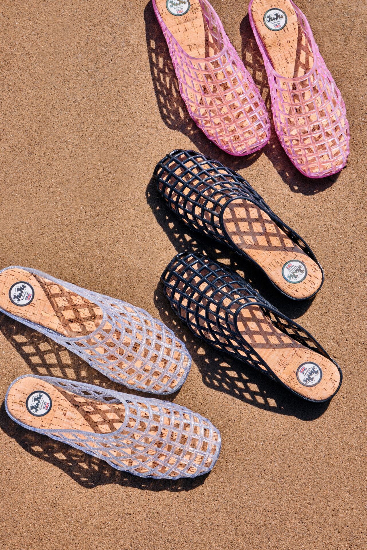 Three pairs of JUJU Jellies Dani Mules, one in pink, one in black, and one in clear, sitting on sand.