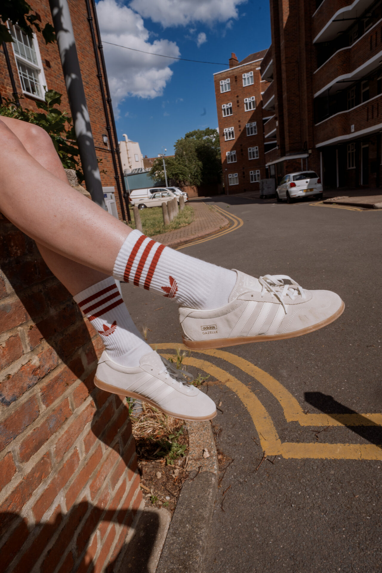 Close up of @biig_piig wearing the adidas Gazelle Lo Pro in off-white with white and red adidas socks, sitting on a brick wall.