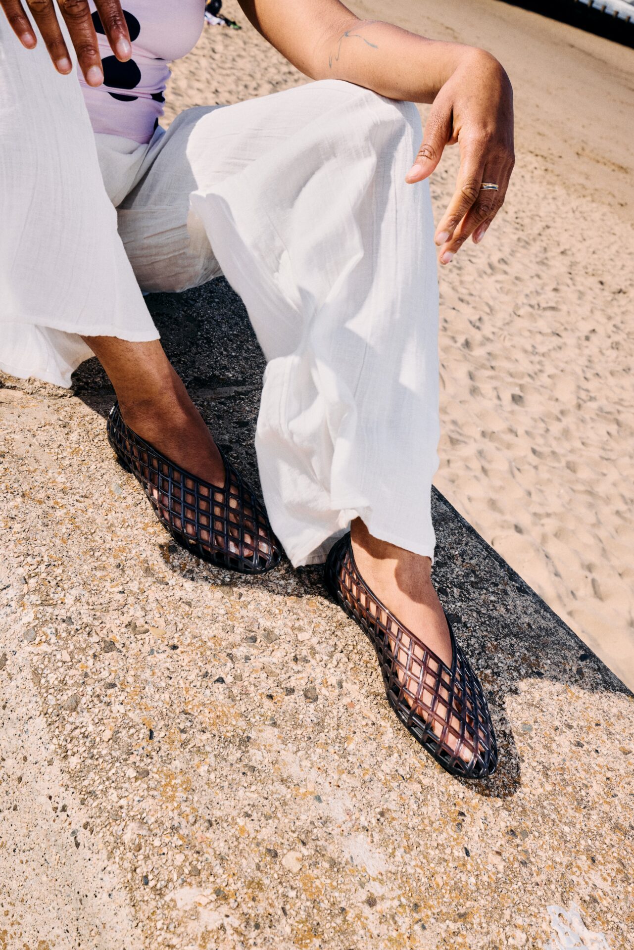 Person wearing the JUJU Jelly Maddi Ballerina in black with flowy white trousers, sitting at a beach.