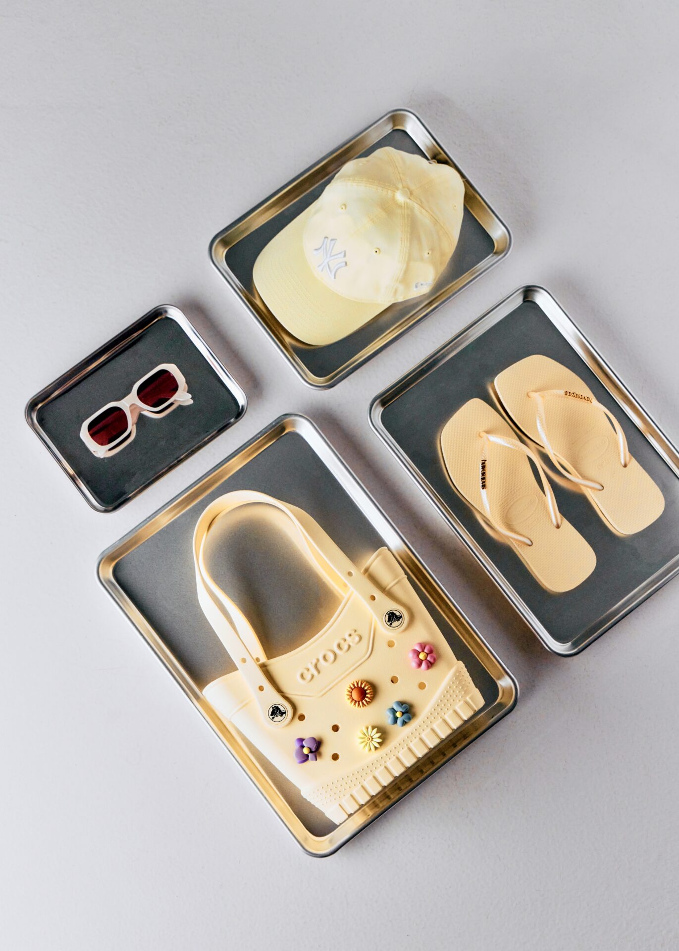 A pair of yellow sunglasses, yellow Havaianas flip flops, a yellow cap, and a yellow Crocs bag each sitting on small metal trays.